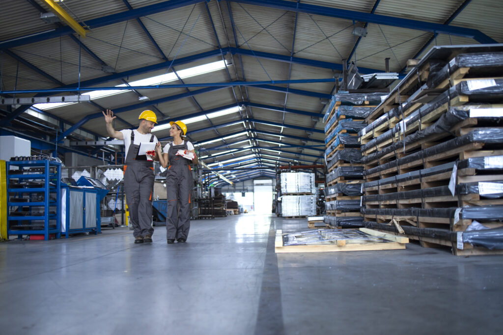 factory workers in work wear and yellow helmets walking through industrial production hall and sharing ideas about organization.